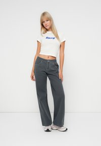 Vit kortärmad T-shirt med blå logotyp, i kombination med löst sittande, mörkgrå jeans. Slät tyg med synlig sömnad och två framfickor.