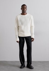Weißer Strickpullover mit strukturiertem Herzmuster und gerippten Bündchen, kombiniert mit schwarzen Hosen mit einem weißen Seitenstreifen und schwarzen Schuhen.