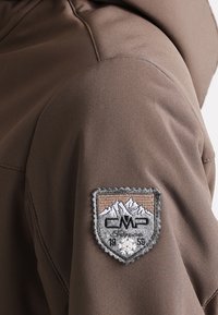 Vêtement d'extérieur marron avec une texture lisse. Présente un patch cousu représentant des montagnes, les lettres « CMP » et un flocon de neige, situé sur l'épaule.