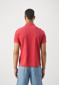 Rdeča polo majica iz bombaža, kratki rokavi, s ovratnikom in bočnimi zarezi, nošena z svetlo modrimi jeans kratkimi hlačami.