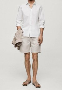 Biała koszula na guziki, jasnobeżowe szorty, brązowe sandały. Trzyma beżową kurtkę. Jasna karnacja, częściowo widoczne nogi. Prosty design.