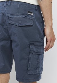 Mørkeblå cargo shorts laget av lett stoff, med sidelommer og en baklomme med svart merke.