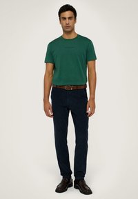 Zelené bavlněné tričko s kulatým výstřihem a jemným logem tištěným na prsou, v kombinaci s tmavě námořnickými tapered kalhotami a hnědými koženými botami.