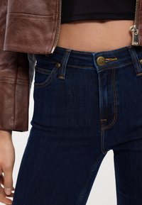 Gros plan d'une personne portant un jean Lee bleu foncé avec des surpiqûres orange et une veste en cuir marron avec une fermeture éclair, montrant le ventre et la main.