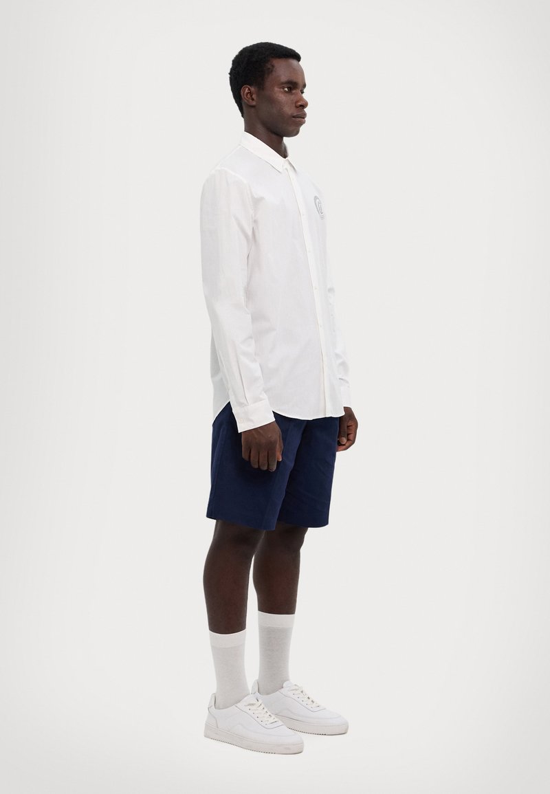 Camicia bianca a maniche lunghe con colletto, chiusura a bottoni e logo sul petto, abbinata a pantaloni corti blu navy e sneakers bianche con calzini leggeri.