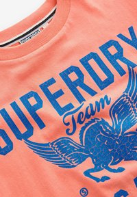 Korallfärgad bomulls t-shirt med djärva, texturerade blå grafiker av "SUPERDRY," "Team," och en vinge design, med ribbad halsringning.