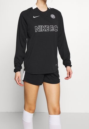 Person trägt ein schwarzes Nike FC Sporttrikot mit langen Ärmeln, schwarze Shorts mit weißem Streifen und weiße knielange Socken vor einem schlichten Hintergrund.