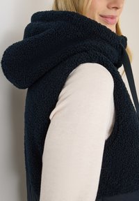 Chaleco de forro polar marino con capucha, que presenta una textura suave y un diseño bicolor, contrastado con una camisa crema más clara debajo.