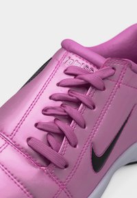 Ροζ αθλητικό παπούτσι Nike με μαύρο σήμα, ροζ κορδόνια και ορατές ραφές στο πάνω ύφασμα, εμφανίζεται σε κοντινή γωνιακή λήψη.