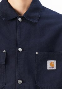 Chaqueta azul marino de algodón con cuello, botones a presión de metal, dos bolsillos en el pecho y un parche con el logo de Carhartt.