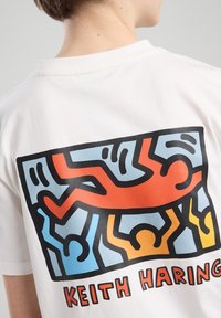 White cotton t-shirt featuring a Keith Haring graphic on the back, depicting colorful abstract figures in blue, orange, and yellow against a black outline.