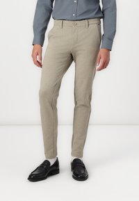 Pantalones de color beige claro en un diseño ajustado, confeccionados con una tela texturada con detalles de costura lateral; combinados con mocasines negros y calcetines grises.