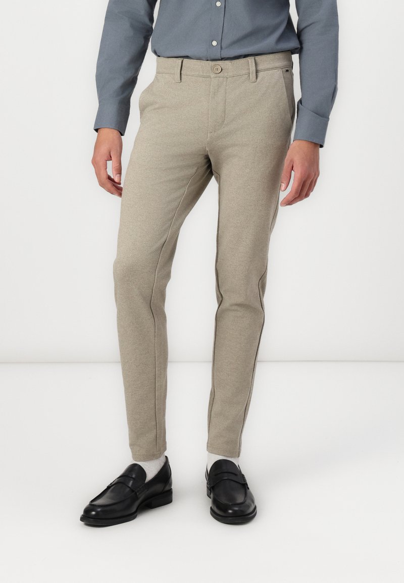 Pantalones de color beige claro en un diseño ajustado, confeccionados con una tela texturada con detalles de costura lateral; combinados con mocasines negros y calcetines grises.