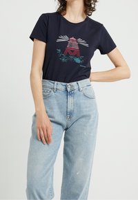 Kobieta w granatowej koszulce z grafiką latarni morskiej w czerwonym i białym kolorze oraz jasnoniebieskich spodniach z wysokim stanem, na jednolitym tle.