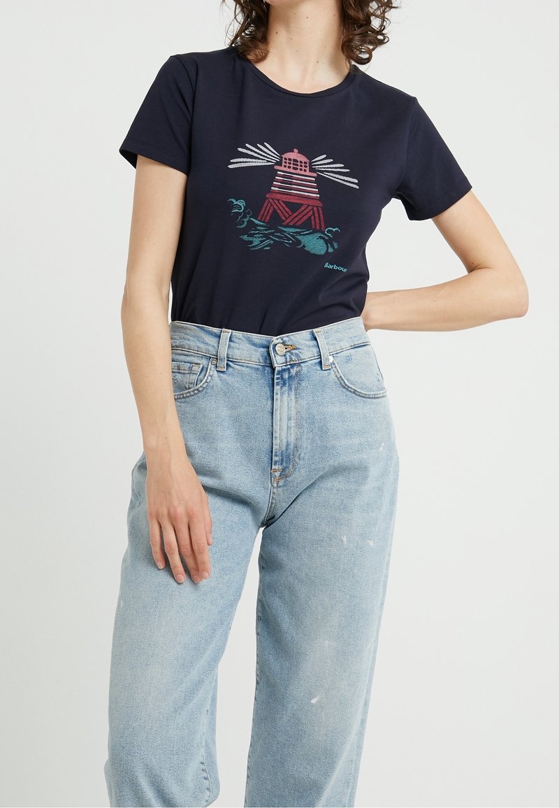 Kobieta w granatowej koszulce z grafiką latarni morskiej w czerwonym i białym kolorze oraz jasnoniebieskich spodniach z wysokim stanem, na jednolitym tle.