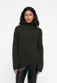 Dunkelgrüner, gerippter Rollkragenpullover mit langen Ärmeln, aus strukturiertem Strickstoff. Tragen mit schwarzen Lederhosen.
