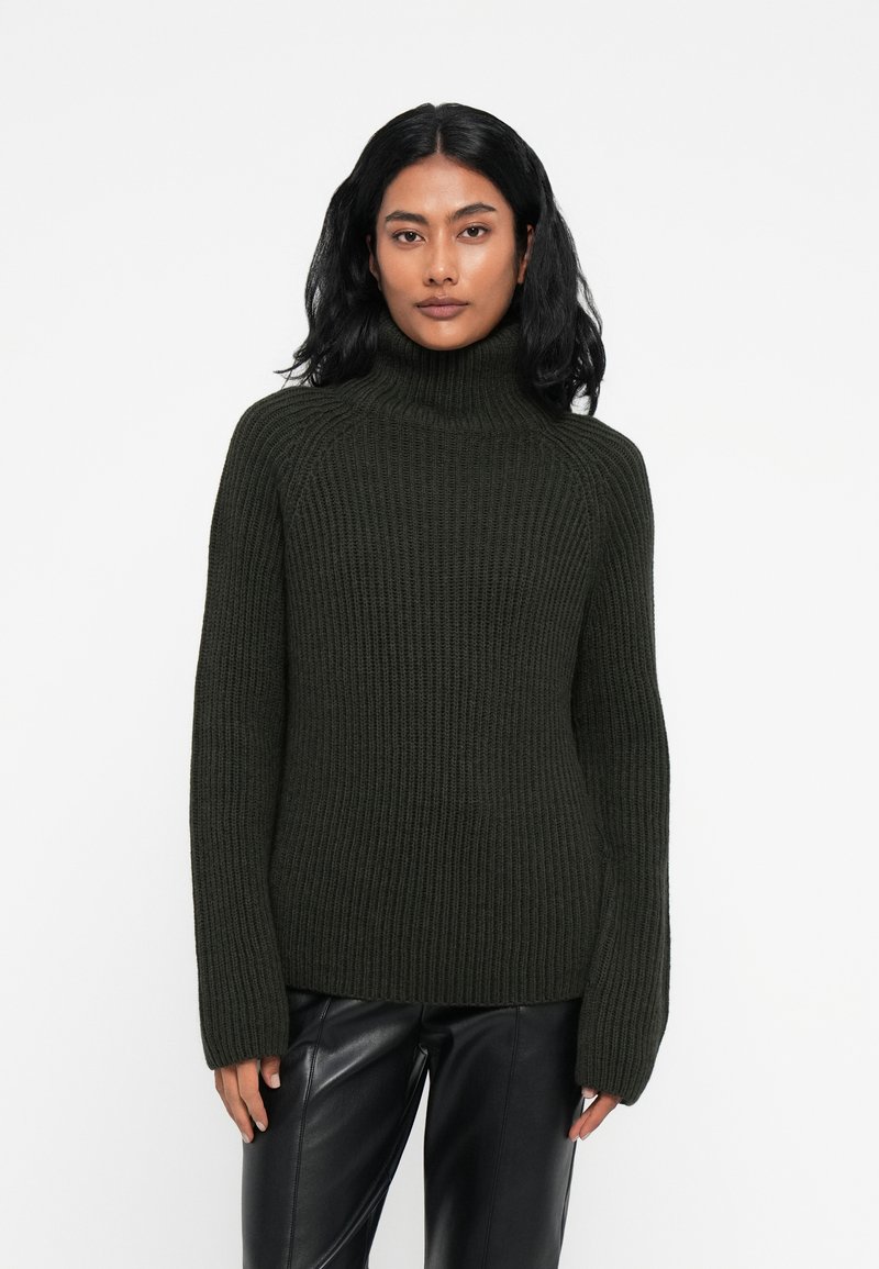Dunkelgrüner, gerippter Rollkragenpullover mit langen Ärmeln, aus strukturiertem Strickstoff. Tragen mit schwarzen Lederhosen.