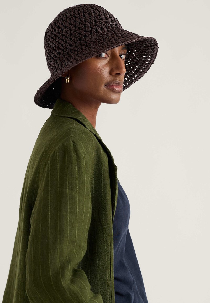 Sieviete, valkājot tumši brūnu austu bļodas cepuri, olīvu zaļas svītrains žaketi un tumši zilas krāsas topu, skatās sāniski pret vienkāršu fonu.