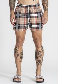 Mann trägt beige, schwarz und rot karierte Shorts mit schwarzer Kordel, passende karierte Sandalen und mehrere Tattoos an Armen und Beinen.