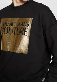 Černá mikina s velkým zlatým fóliovým logem "VERSACE JEANS COUTURE", s žebrovaným výstřihem a měkkou texturou.