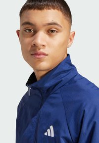 Mörkblå zip-jacka med hög krage, som har en vit logotyp på vänster sida och en slät, lätt tygtextur.