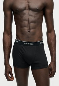 Buste masculin musclé à la peau foncée portant un boxer Calvin Klein noir avec logo sur la ceinture, debout devant un fond clair uni.