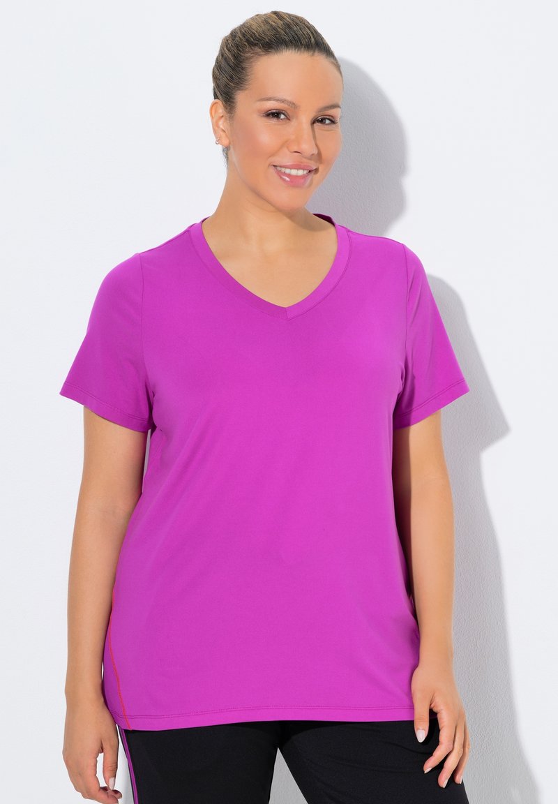 Femme aux cheveux attachés portant un t-shirt violet vif à col en V et manches courtes, et un pantalon noir, souriante, se tenant devant un fond blanc.