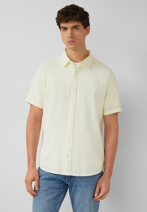 Junger Mann mit kurzärmeligem, hellcremefarbenem, texturiertem Hemd mit Knöpfen und blauer Jeans, der vor einem schlichten grauen Hintergrund steht.