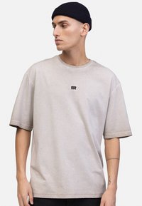 Lys beige t-skjorte med korte ermer og rund hals, samt en avslappet passform. Lite svart logo på brystet. Glatt tekstur, minimalistisk design.