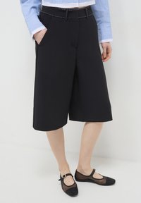Persona che indossa pantaloni corti neri fino al ginocchio, una camicia a maniche lunghe a righe blu e scarpe Mary Jane nere trasparenti in piedi su un pavimento bianco.
