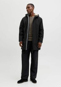 Czarna wodoodporna kurtka z kapturem, wyłożona miękkim beżowym materiałem, w połączeniu z jasnoszarym swetrem i ciemnymi spodniami.