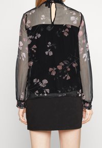 Blouse noire avec un motif floral transparent agrémenté d'accents roses, manches longues avec poignets froncés et un détail dos avec une ouverture en forme de goutte.