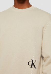Sweatshirt beige à col rond en coton, avec un logo noir "CK" et du texte blanc le long de la manche. Texture lisse, coupe décontractée.