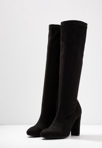 Bottes noires à talons hauts, montantes jusqu'à la cuisse, fabriquées en tissu doux, présentant un design élégant, une texture lisse et un bout arrondi.
