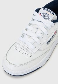 Hvit Reebok Classic-sneaker med perforert tå, polstret marineblå krage, hvite lisser og Reebok-logo med Union Jack på siden.