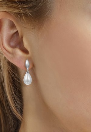 Pendiente de botón plateado con forma de gota y colgante de perla en forma de lágrima, llevado en la oreja izquierda de una mujer con piel clara y cabello rubio.