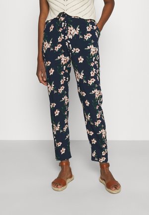 Person trägt marineblaue Blumenhose und braune Sandalen mit Knöchelriemen und steht mit den Händen in den Taschen vor einem schlichten Hintergrund.