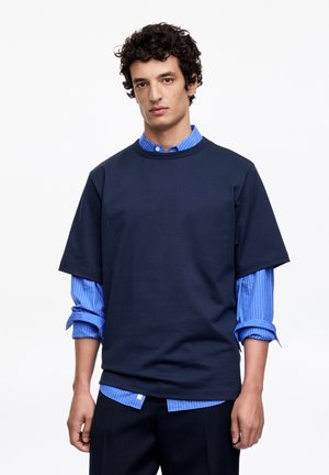 Hombre con cabello rizado que lleva una camisa azul marino de manga corta y holgada, superpuesta sobre una camisa de botones azul a rayas, posando frente a un fondo blanco.