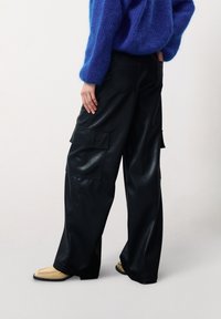 Pantalons cargo en cuir synthétique noir avec une coupe large, dotés de plusieurs poches latérales et d'un éclat lisse, associés à des chaussures beiges.