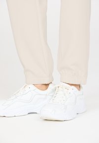 Beige tapered Jogginghosen mit elastischen Bündchen, kombiniert mit weißen Sportschuhen, die über strukturierte Muster und hellgraue Akzente verfügen.
