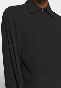 Camisa negra de manga larga hecha de tejido liviano, con un cuello clásico, detalle de botones y suaves pliegues a lo largo de las mangas.