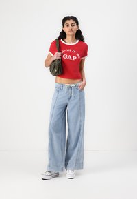 Punase lühike T-särk valge kaeluse ja "GAP" logoga, koos helesiniste laia säärega teksade ja valgete spordijalatsitega. Oliivroheline õlakott.