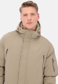 Giacca imbottita beige con collo alto, tasche con zip laterali e sul petto, e cappuccio con coulisse regolabile. Finitura del tessuto liscia.