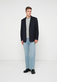 Blazer blu navy con rever a punta, due tasche frontali, abbinato a una maglietta a righe grigie e bianche, jeans a gamba larga azzurri chiari e scarpe nere.