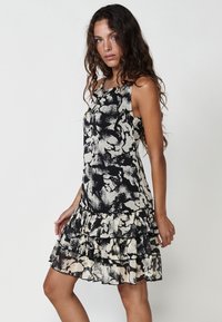 Ärmelloses Kleid mit floralem Muster in Schwarz und Creme, mit Rüsche am Saum und leichtem Stoff mit transparenten Details.