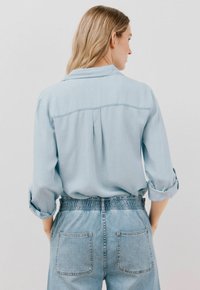 Camicia azzurro chiaro a maniche lunghe con colletto, dettaglio con piega sul retro e maniche arrotolate, abbinata a pantaloni in denim azzurro chiaro con vita elasticizzata.