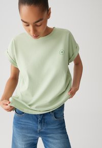 Maglietta verde chiaro a maniche corte con risvolti e un piccolo dettaglio grafico circolare sul petto. Texture morbida, vestibilità comoda.