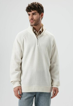 White zip-up pullover made of textured fabric, featuring a high collar, ribbed cuffs, and a relaxed fit. Worn over a light-colored shirt.