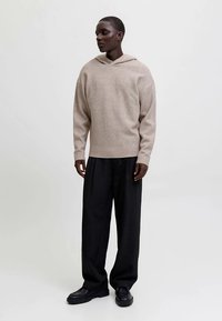 Sweat à capuche en laine beige avec une coupe décontractée et un large décolleté, associé à un pantalon noir à fines rayures. Des chaussures à semelles en caoutchouc complètent le look.