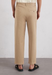 Pantaloni beige con gamba dritta e una tasca posteriore, realizzati in tessuto leggero, abbinati a sandali neri a scivolo. Design minimalista.